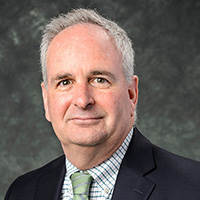 Headshot of David Murphy wearing a dark colored suit and green tie on a dark grey background.